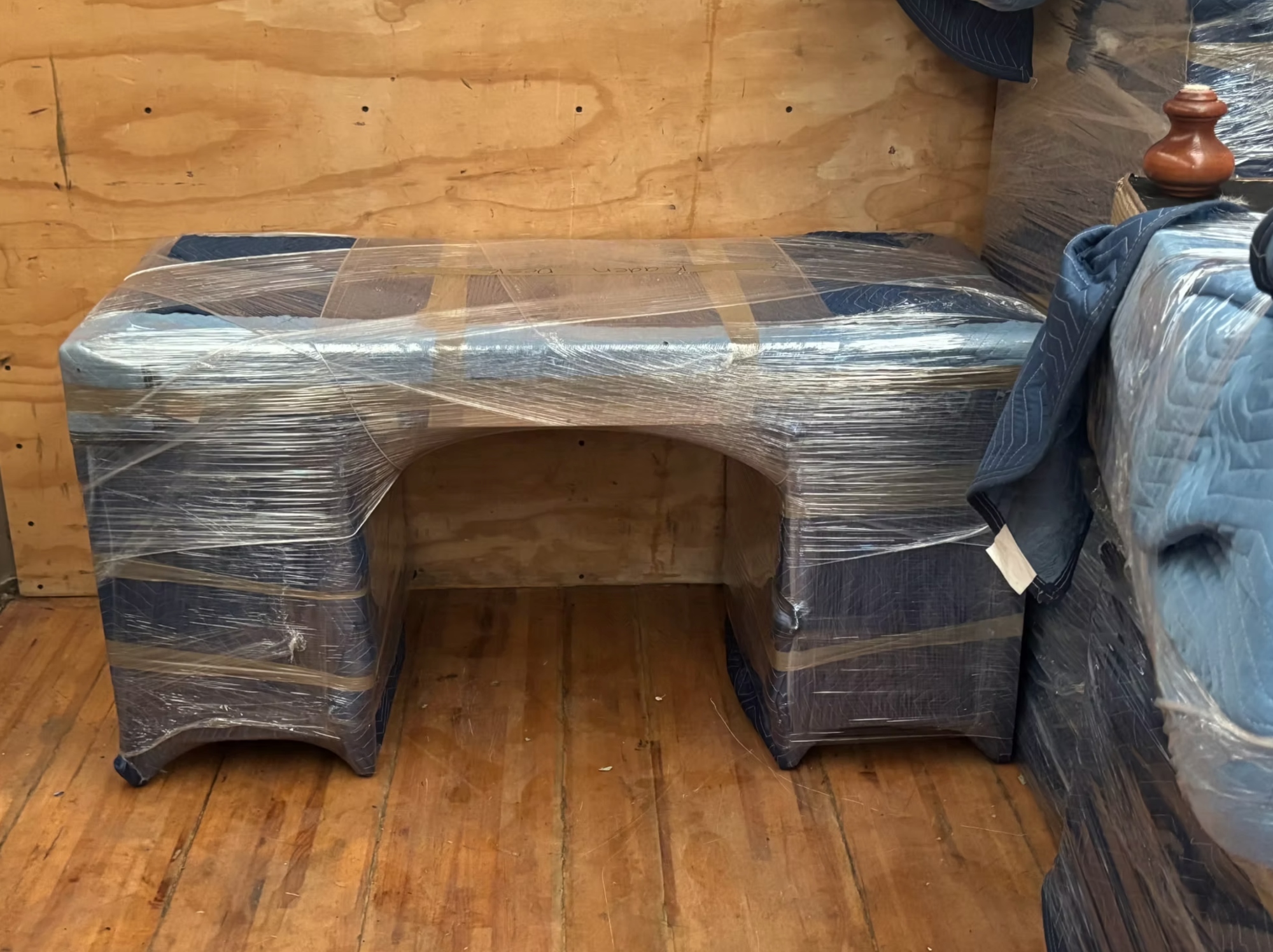 A shrink-wrapped and blanket-padded desk staged beside a wrapped mattress inside the wood-lined moving truck.