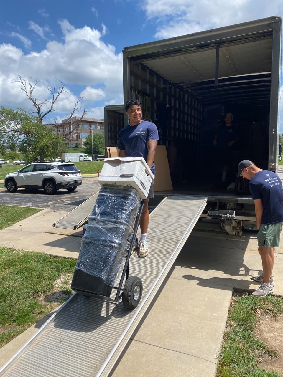 Smiling Box N Haul mover wheeling a shrink-wrapped item topped with a Bankers Box up the truck ramp on a sunny day while a teammate assists.