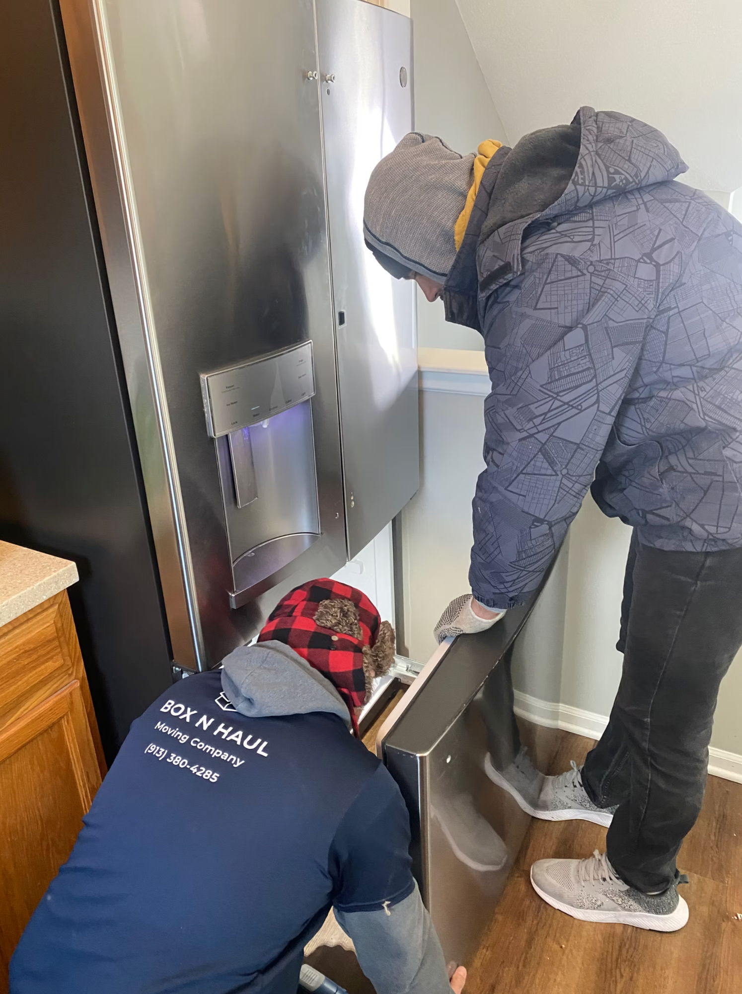 Two Box N Haul movers carefully installing and connecting a stainless steel refrigerator in a kitchen.