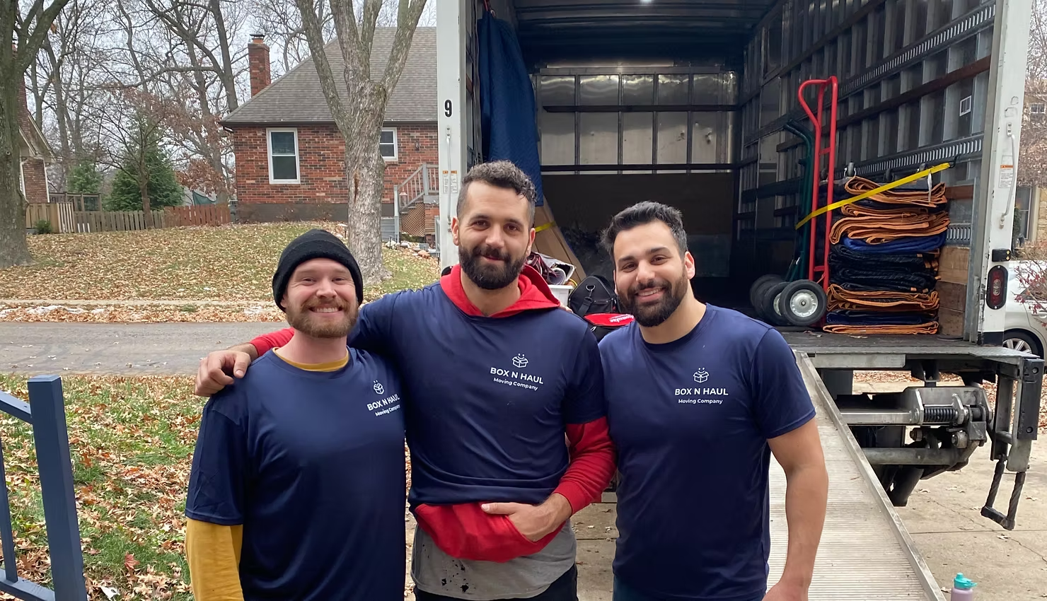 Three smiling Box N Haul movers in branded shirts in front of a loaded moving truck