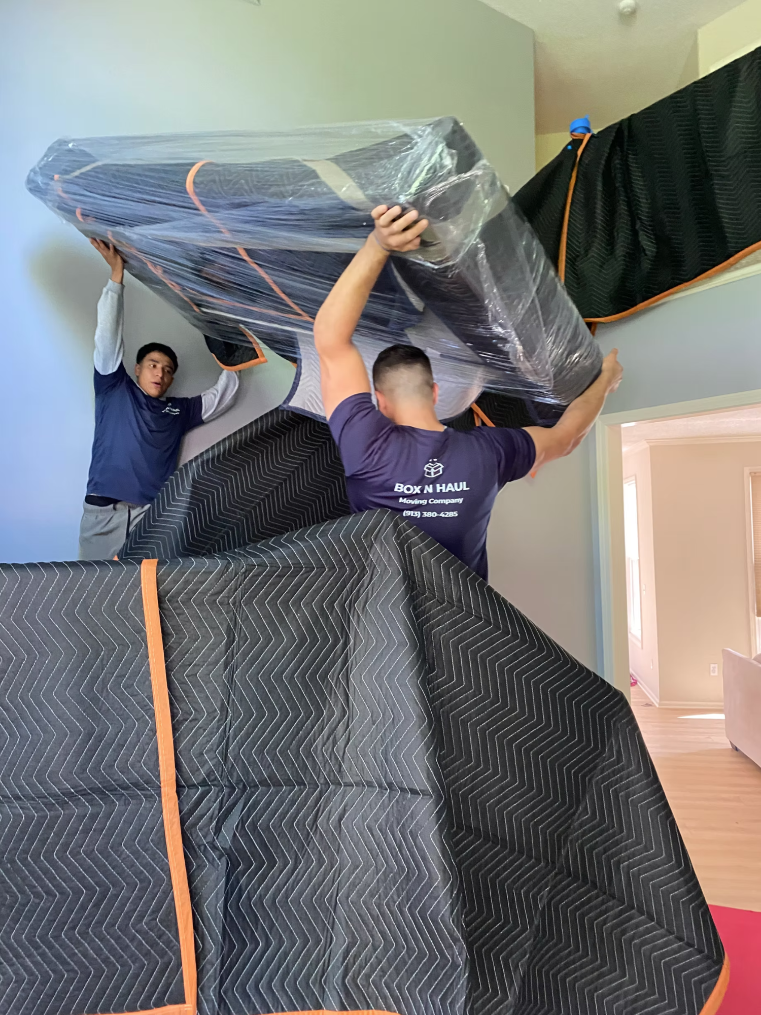 Two Box N Haul movers lifting a large shrink-wrapped, blanket-padded piece of furniture inside a home.