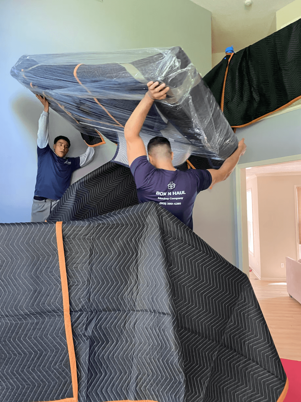 Two Box N Haul movers lifting a large blanket-padded and wrapped piece of furniture inside a home