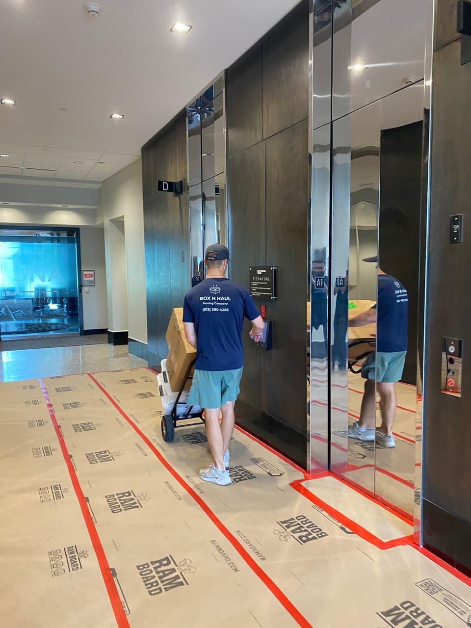 A Box N Haul mover in a branded navy shirt wheeling boxes on a hand truck through a protected office lobby toward the elevators