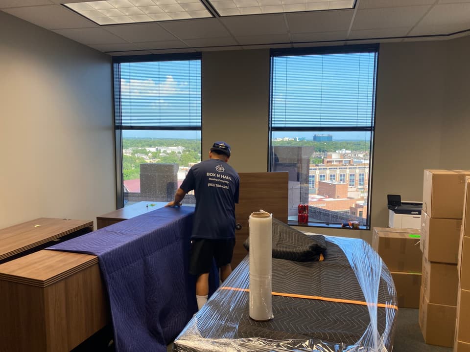 Box N Haul mover wrapping office furniture in moving blankets in a high-rise office with a Kansas City skyline view.