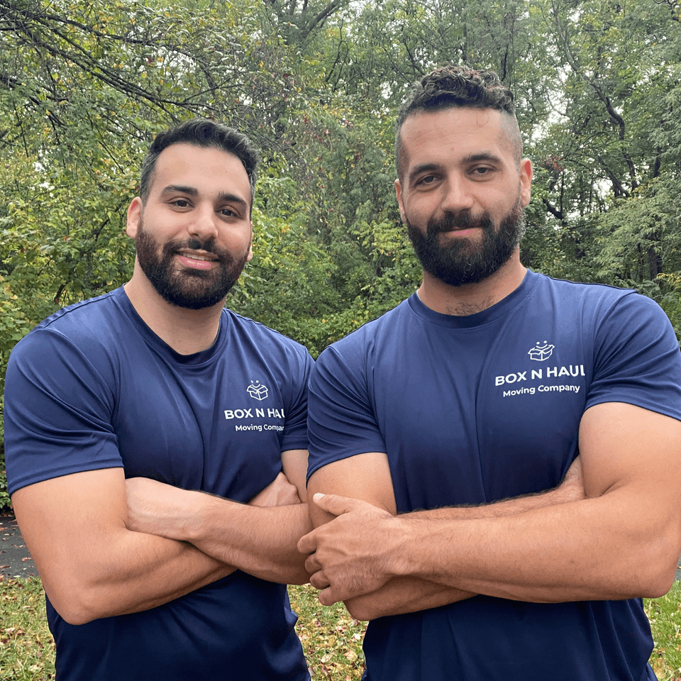 Cam and Kyle of Box N Haul Moving standing outdoors with arms crossed in their navy Box N Haul Moving Company shirts.