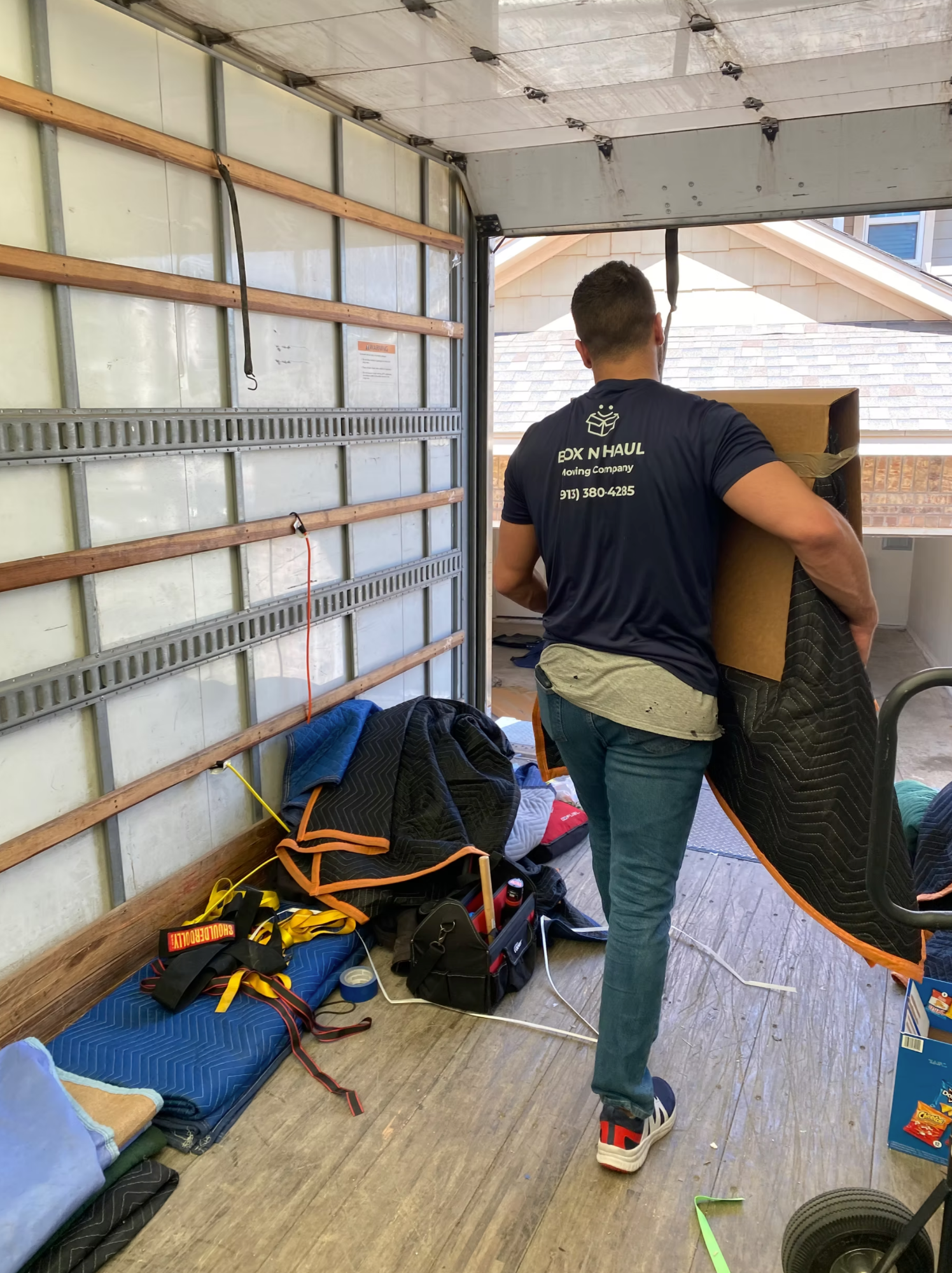 Box N Haul mover in a branded shirt loading a blanket-wrapped item into the back of the moving truck, padding blankets staged on the floor.