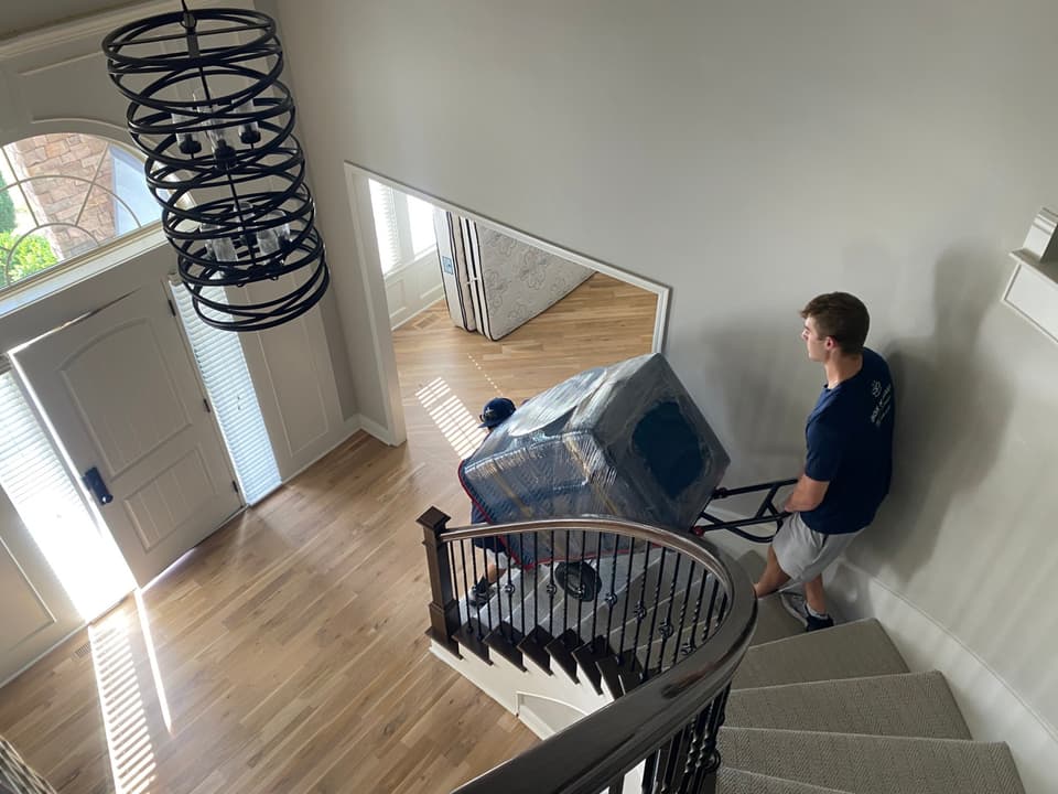 Two Box N Haul movers wheeling a shrink-wrapped, blanket-padded item down a curved staircase on a hand truck.