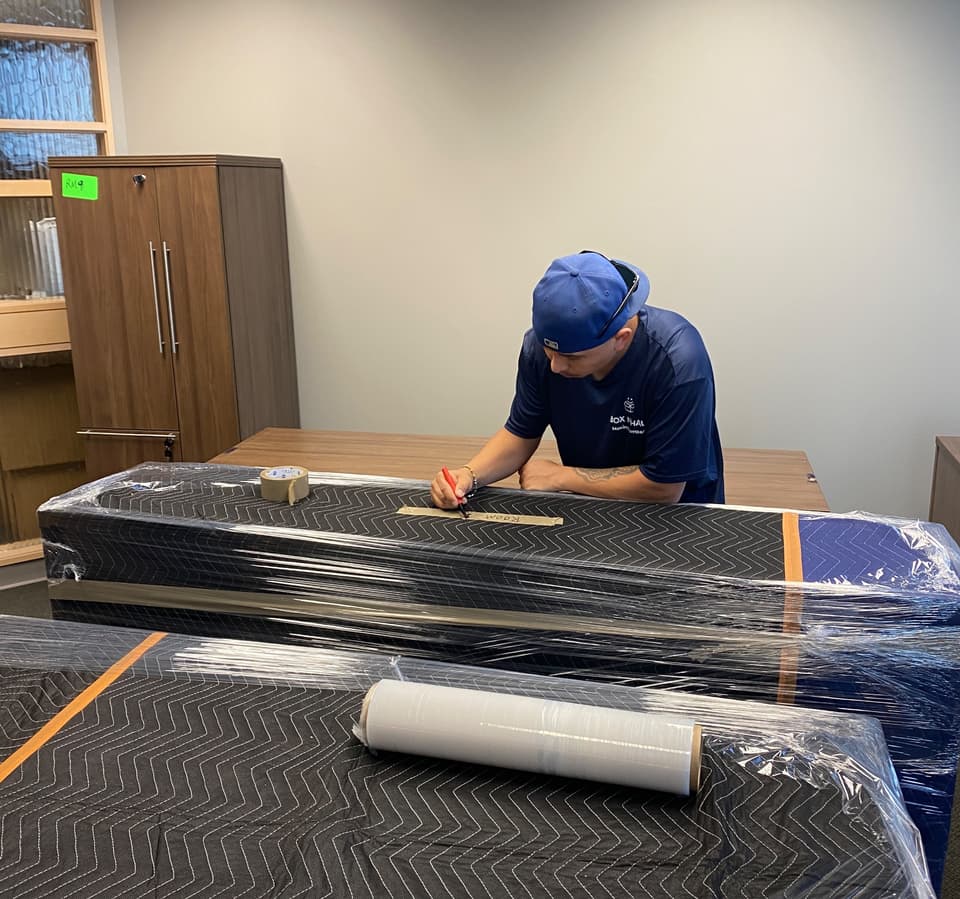 Box N Haul mover in a blue cap labeling a shrink-wrapped, blanket-padded piece of office furniture with a marker.