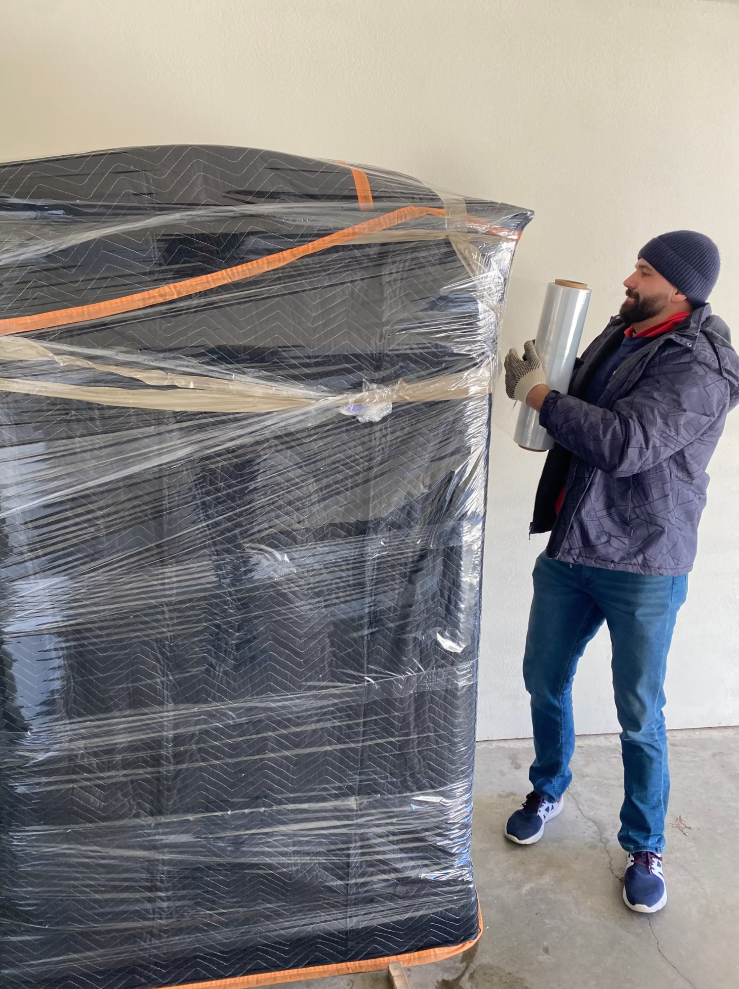 Box N Haul mover in a beanie shrink-wrapping a tall, blanket-padded piece of furniture in a garage.