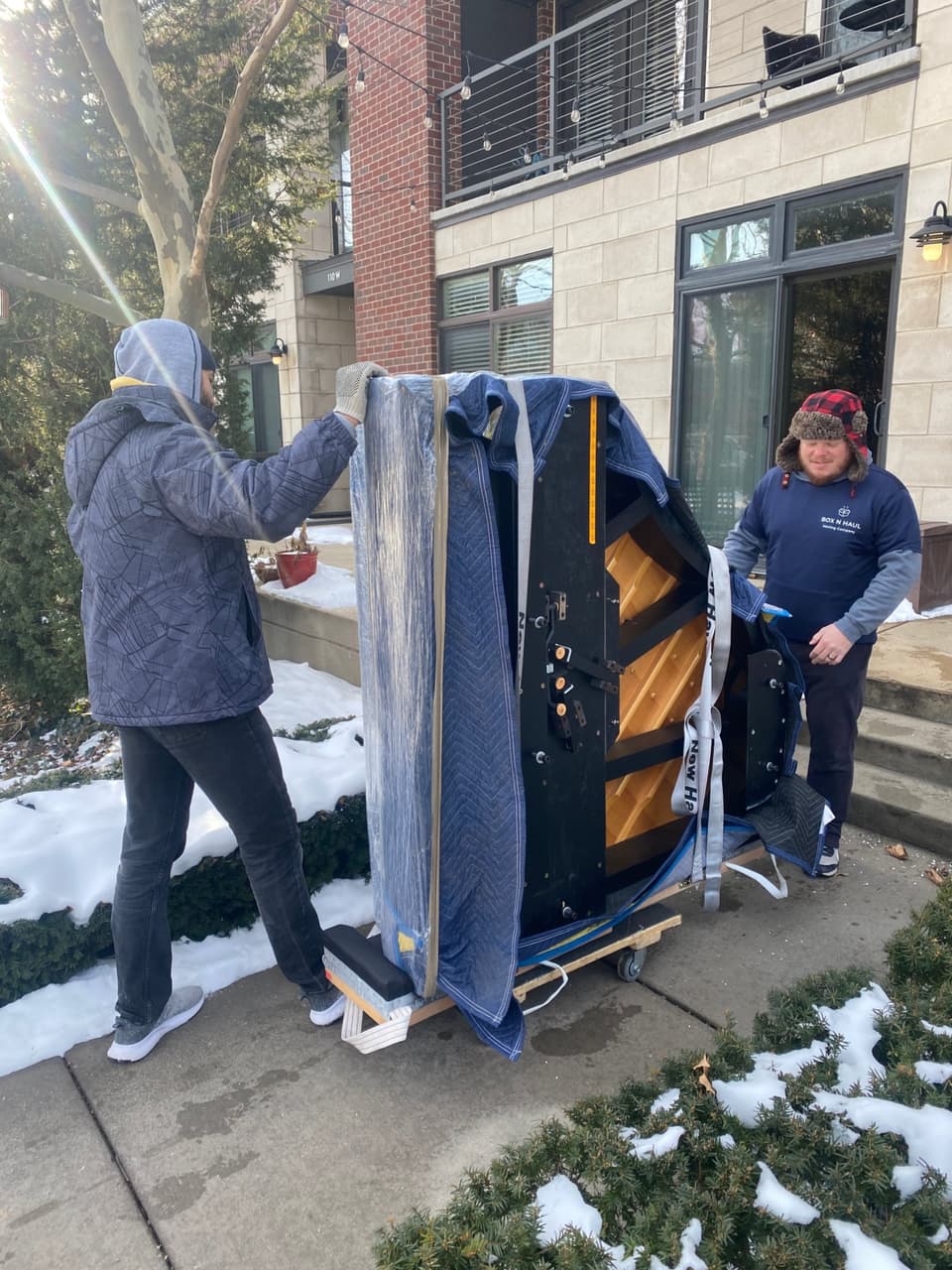 Two Box N Haul movers transporting a blanket-wrapped upright piano on a dolly along a winter walkway