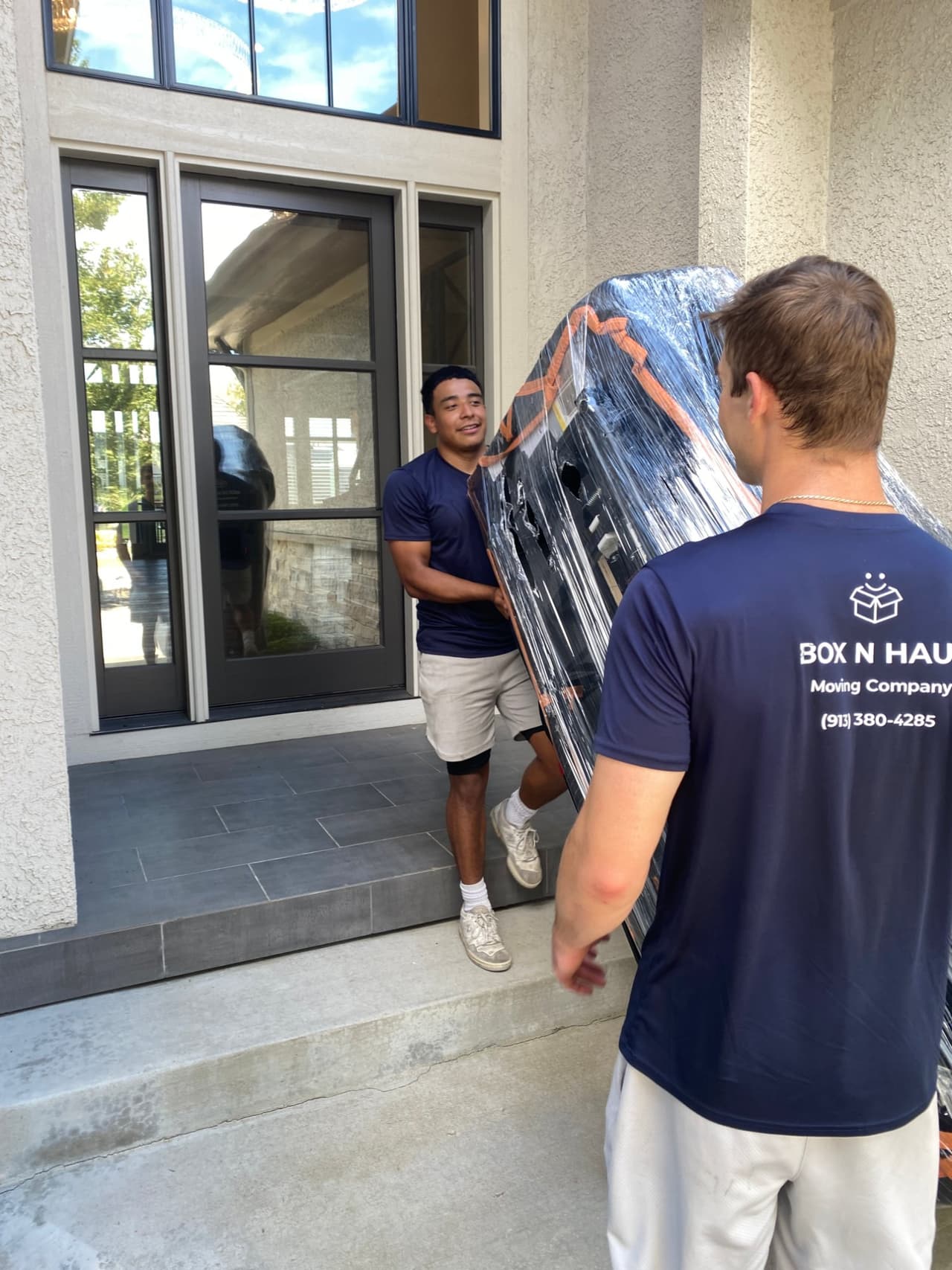 Two Box N Haul movers carrying a shrink-wrapped item through a glass front door