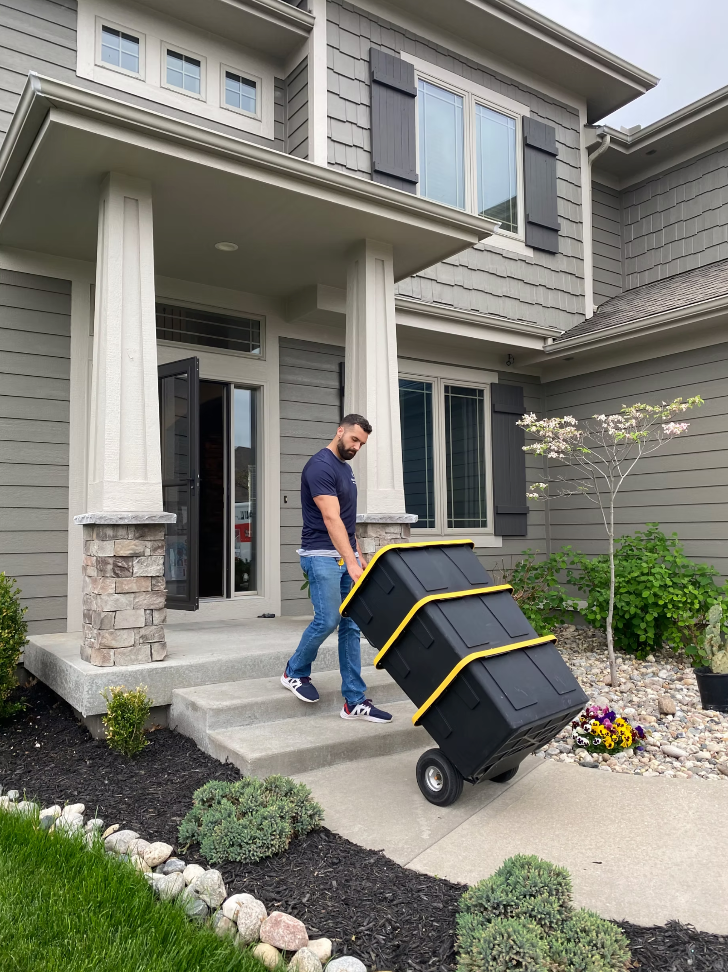 Box N Haul mover wheeling a stack of yellow-trimmed black moving totes on a hand truck down the front steps of a home.