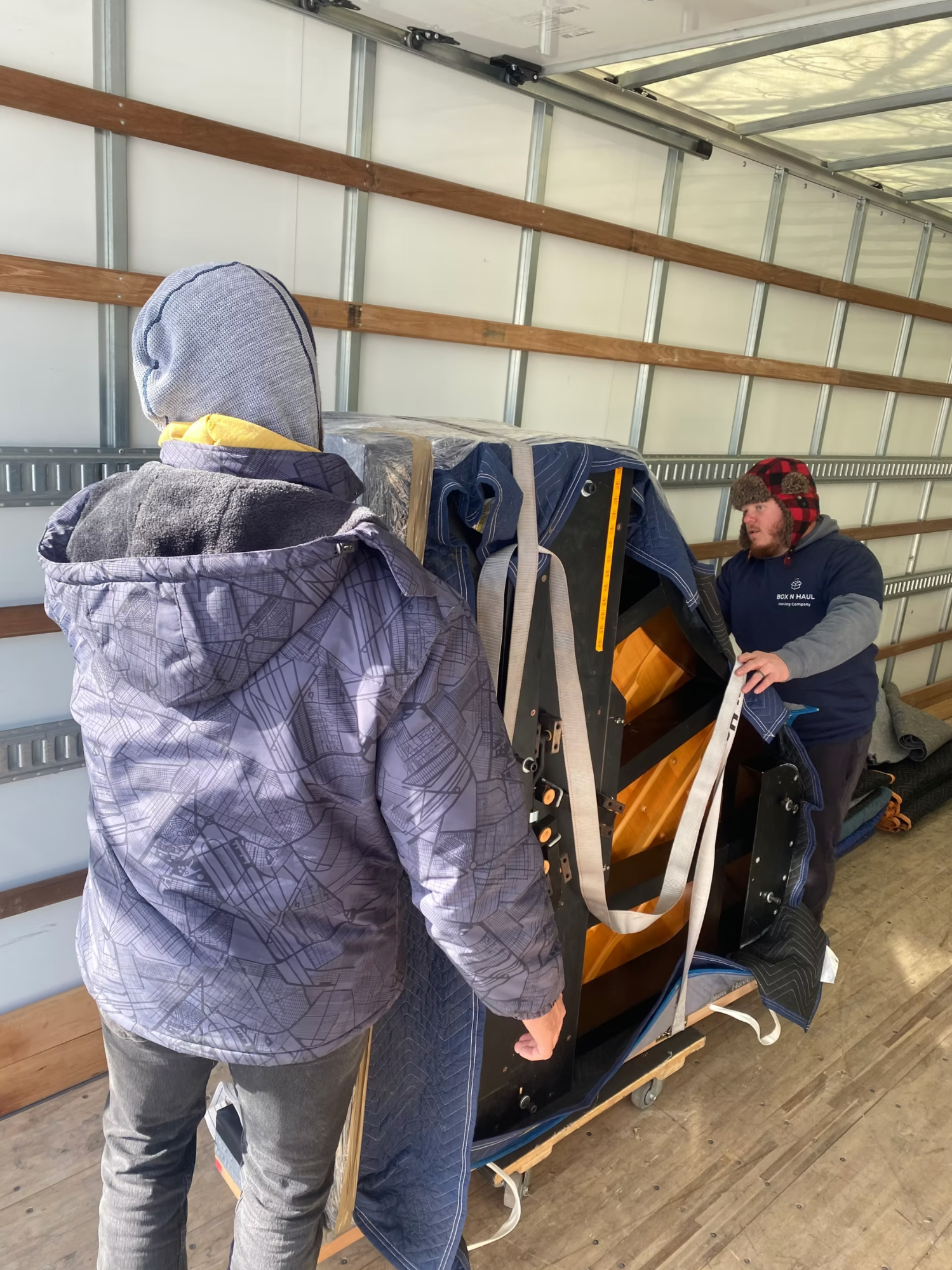 Two bundled-up Box N Haul movers strapping a blanket-wrapped upright piano onto a dolly inside the moving truck on a cold day.