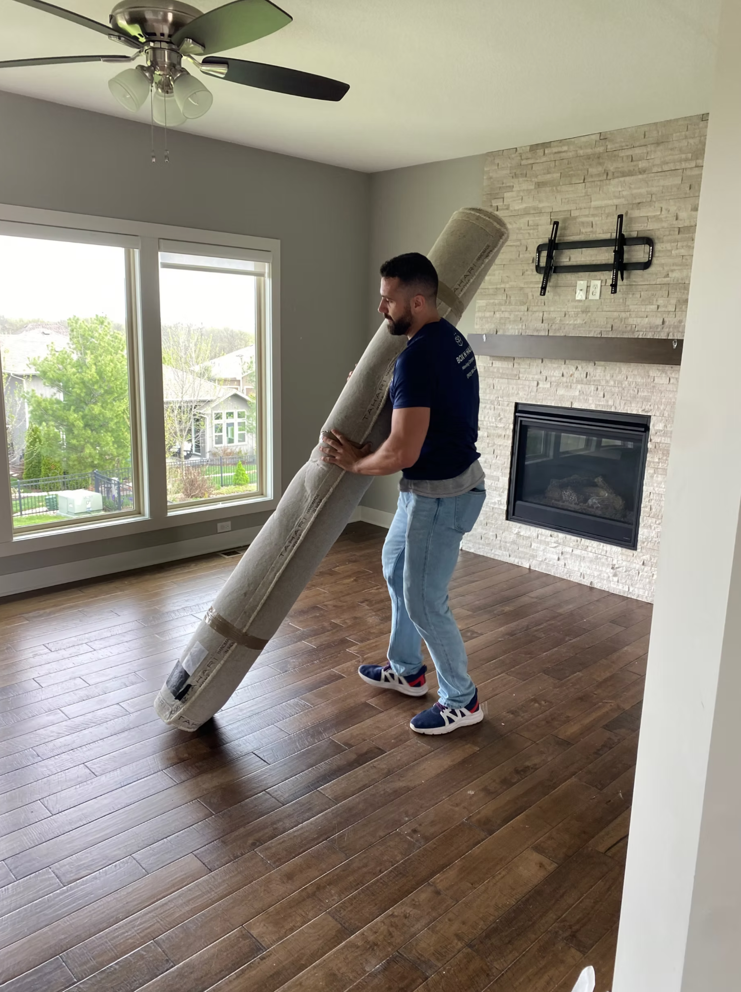 Box N Haul mover carrying a rolled area rug on his shoulder through a living room with a stone fireplace.