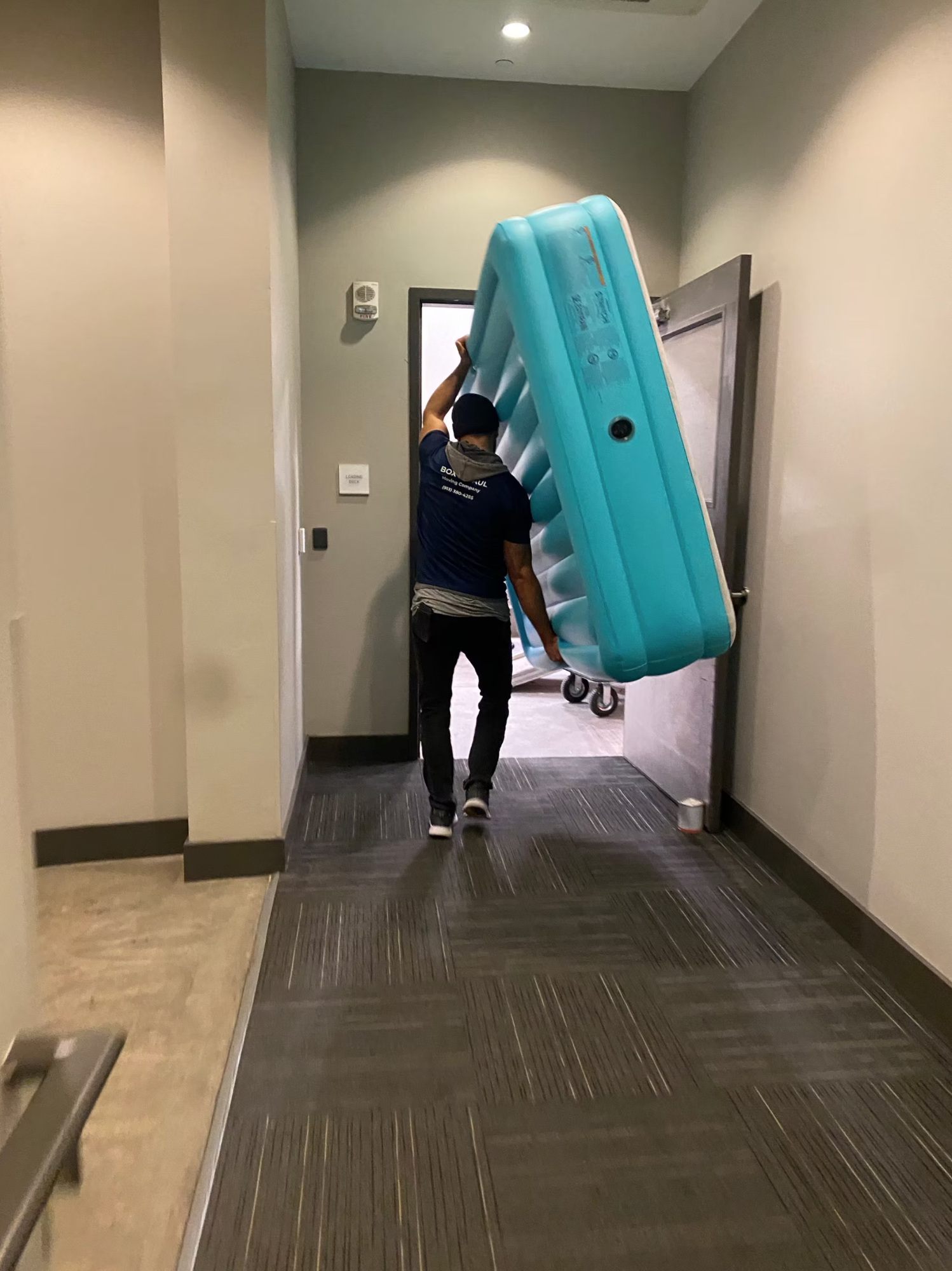 Box N Haul mover wheeling a wrapped inflatable hot tub on a dolly through an office hallway doorway.