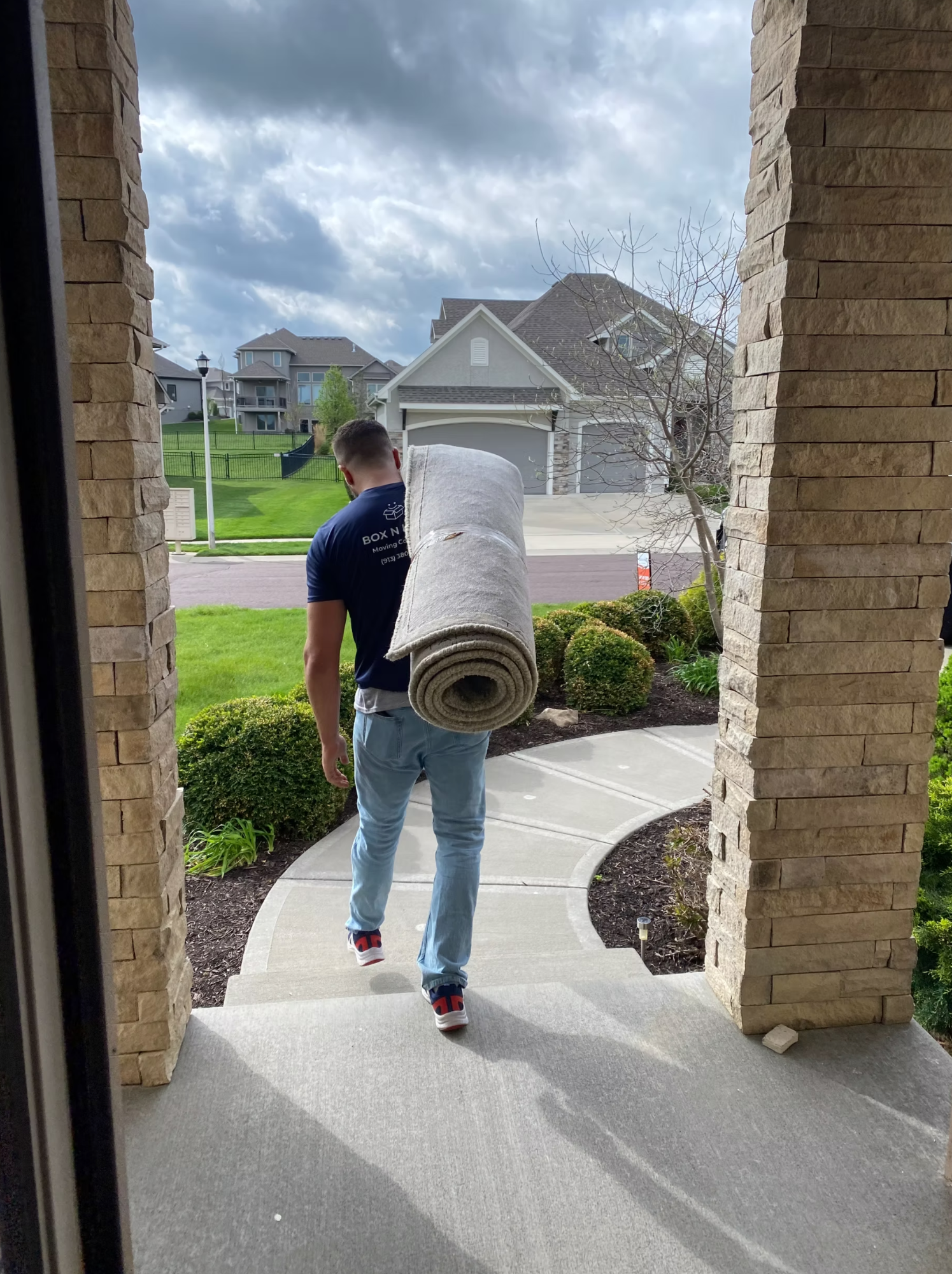 Box N Haul mover in a branded shirt carrying a rolled area rug on his shoulder out the front door toward the walkway.