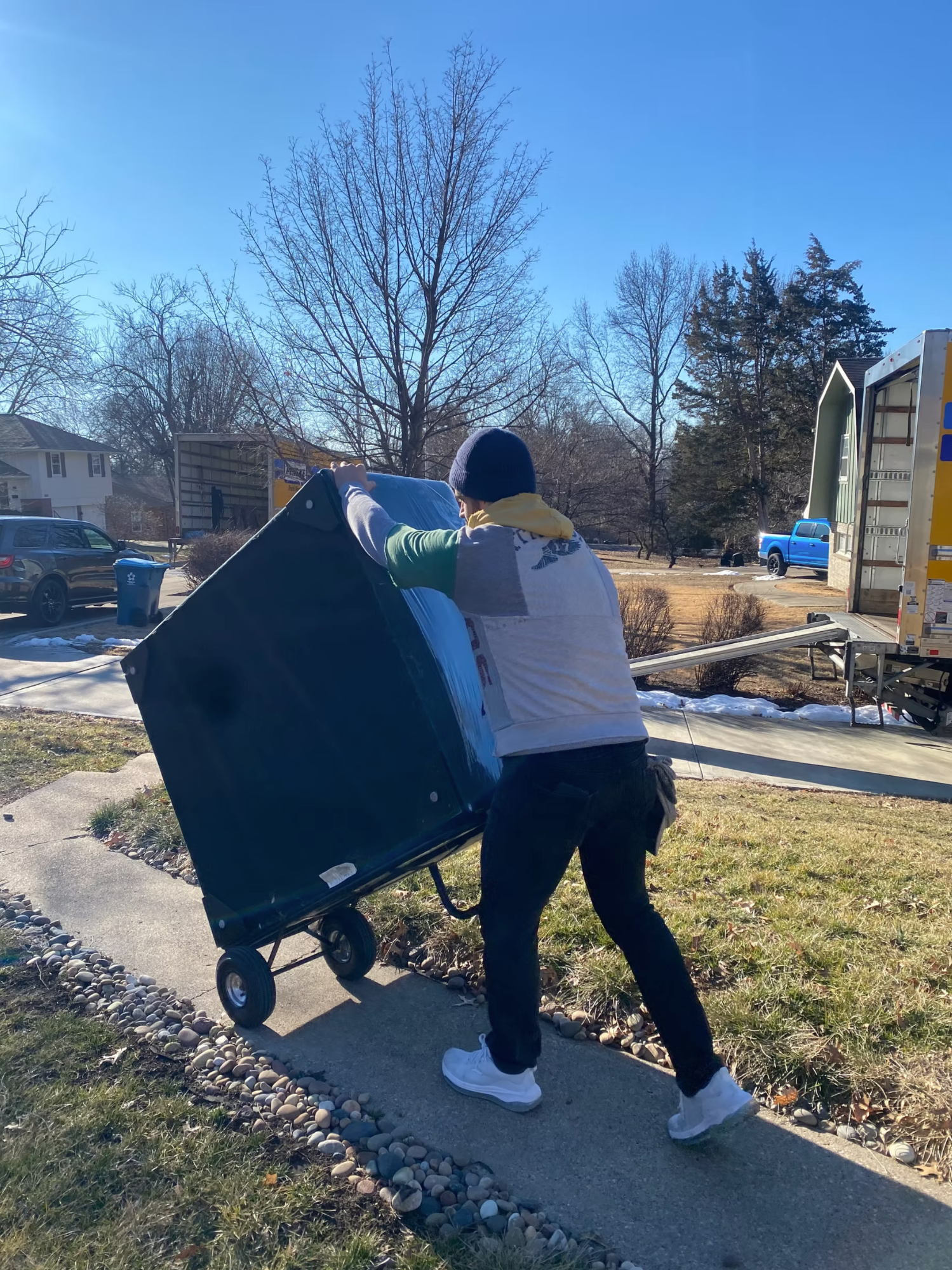 Box N Haul mover wheeling a tall shrink-wrapped item on a dolly up a walkway on a sunny winter day, moving truck parked at the curb.